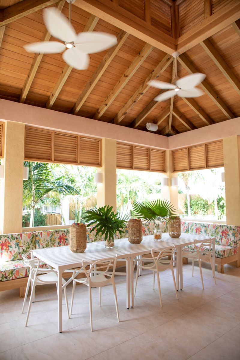 A covered patio in Harbour Island with a cypress beamed ceiling over a long white dining table and parrot print banquette.