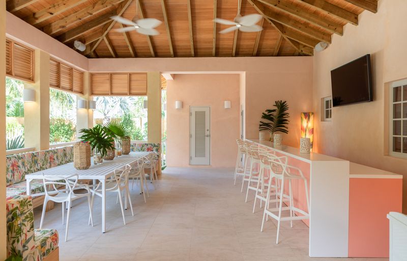 A covered patio in Harbour Island with a pink stucco bar under an open cedar ceiling and white stools.