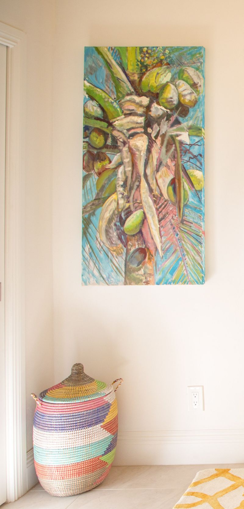 An entry in Harbour Island with a coconut palm painting on a white wall above a lidded woven basket.