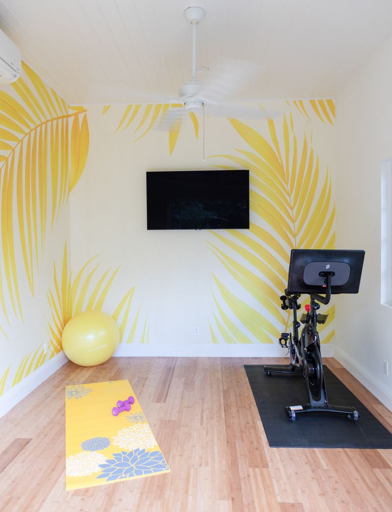 A home gym in Harbour Island with a yellow palm leaf mural and a spin bike on light wood floors.