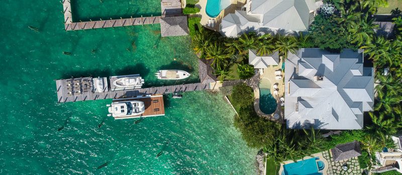 An aerial view of a waterfront property in Harbour Island with wooden docks, moored boats and metal-roofed houses set among palms.