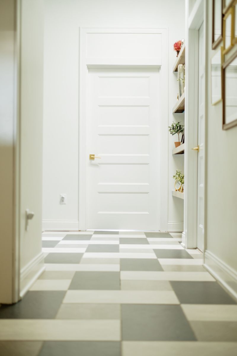 A white hallway in Johnston with a gray and cream checkerboard floor leading to a paneled door with a brass lever.