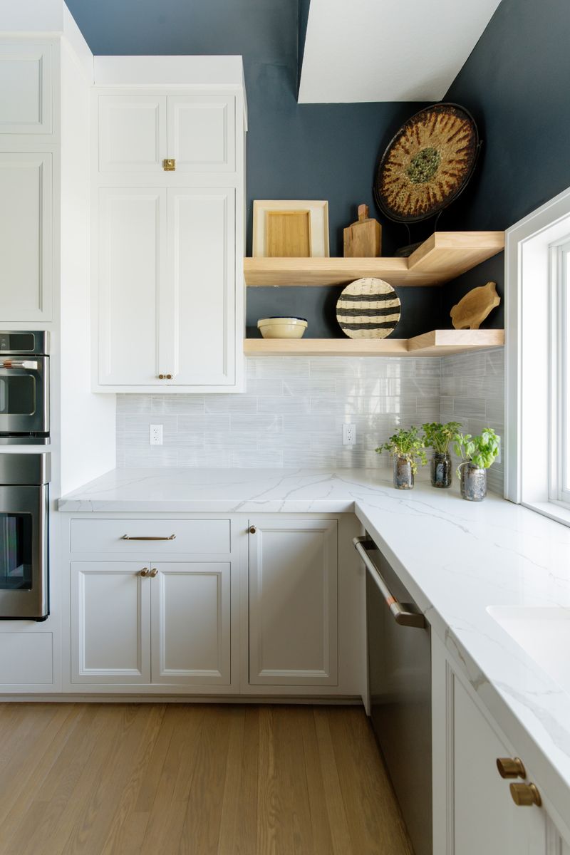 A white kitchen in Johnston with light oak floating shelves on a navy wall above marbled quartz counters.