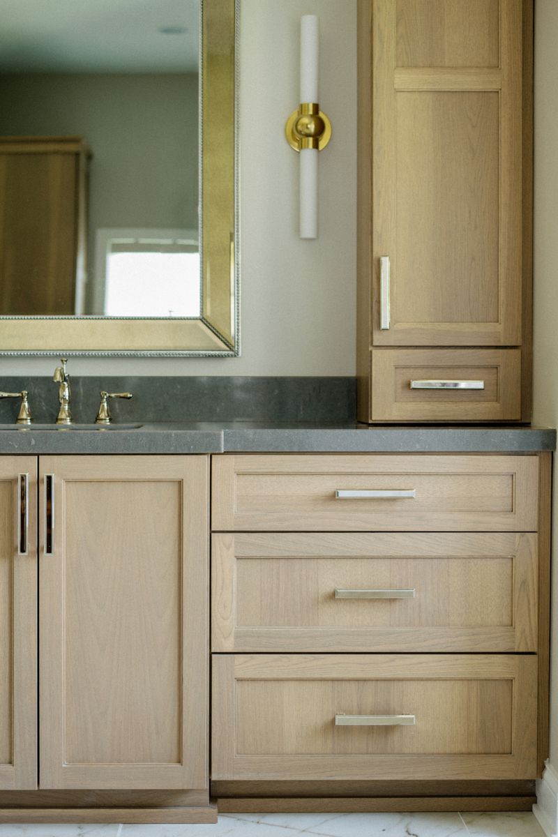 A primary bath in Johnston with a white oak vanity under a honed gray stone counter and a brass sconce.