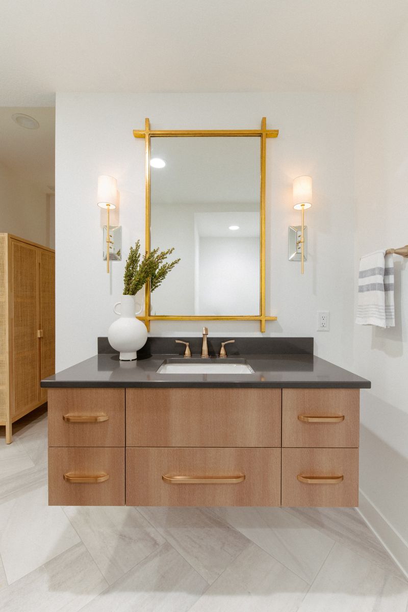 A primary bath in West Des Moines with a floating white oak vanity under a gold framed mirror, topped in charcoal quartz.