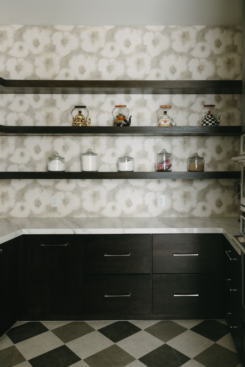 A butlers pantry in West Des Moines with dark wood floating shelves over soft floral wallpaper and marble counters.