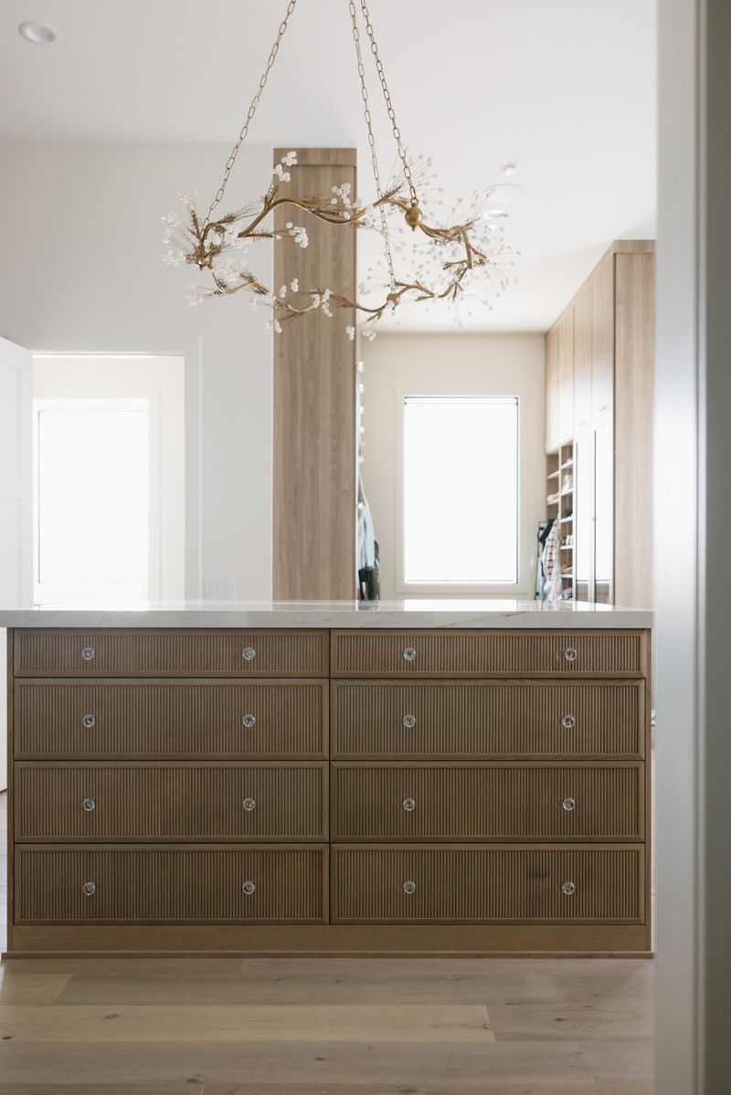 A walk in closet in West Des Moines with a fluted oak drawer island under a branching brass chandelier.