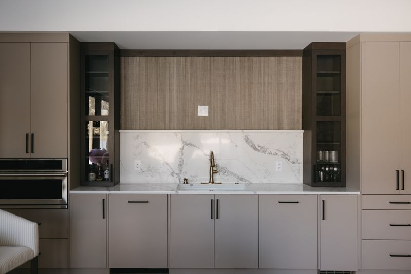 A wet bar in West Des Moines with a brass faucet against a white marble backsplash and greige cabinets.