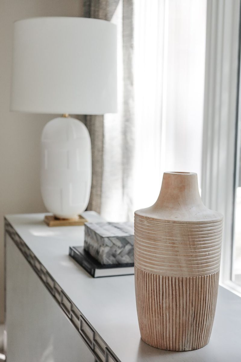 A ribbed carved wood vase on a white entry console in Clive, beside a white ceramic lamp on a brass base.