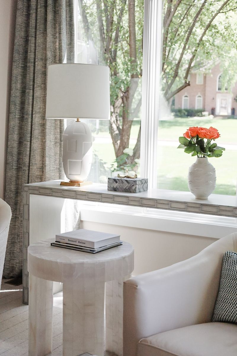 A living room in Clive with a white ceramic lamp on a carved gray console table by the window.
