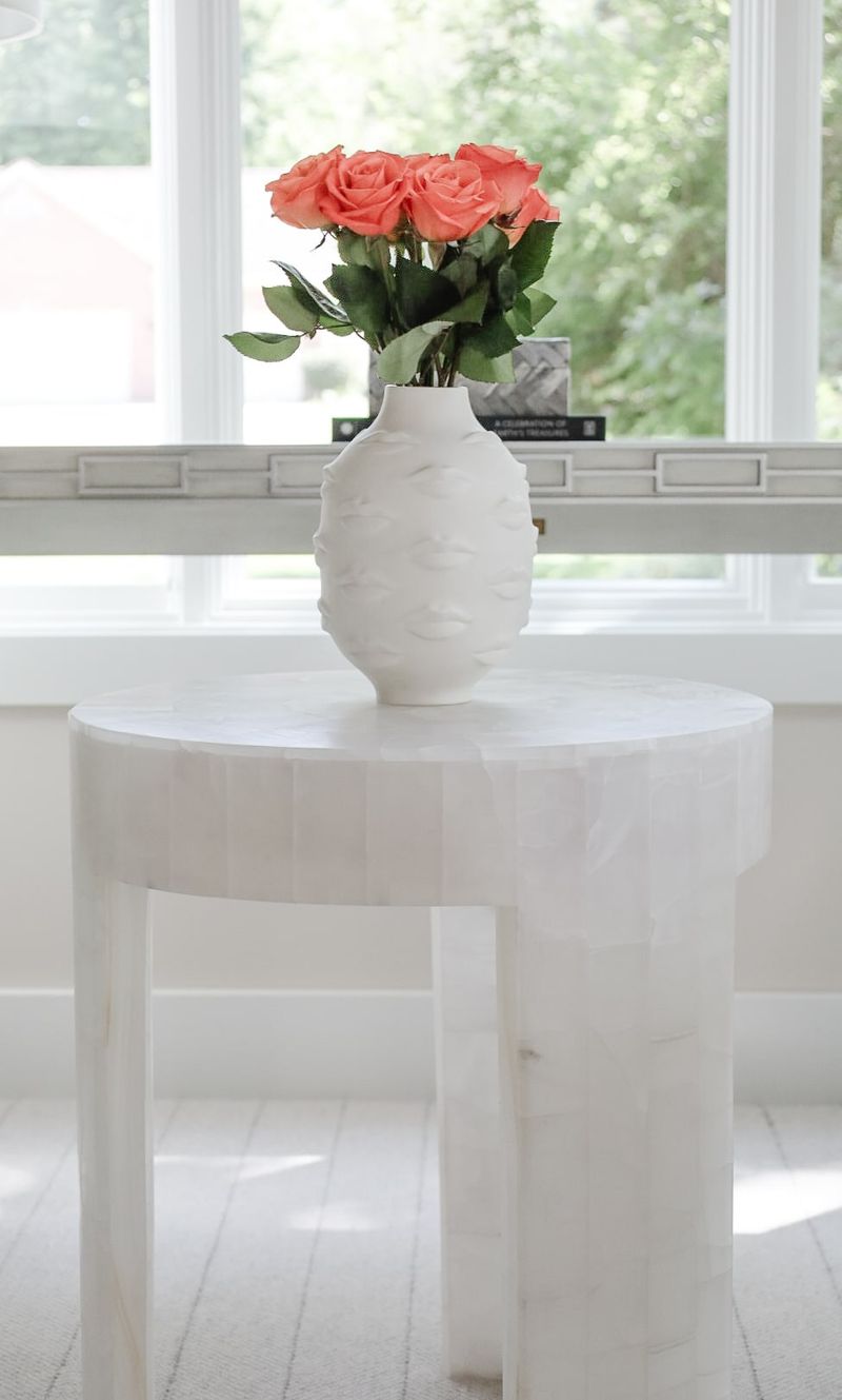 A living room in Clive with a white sculpted face vase on a round stone side table by the window.