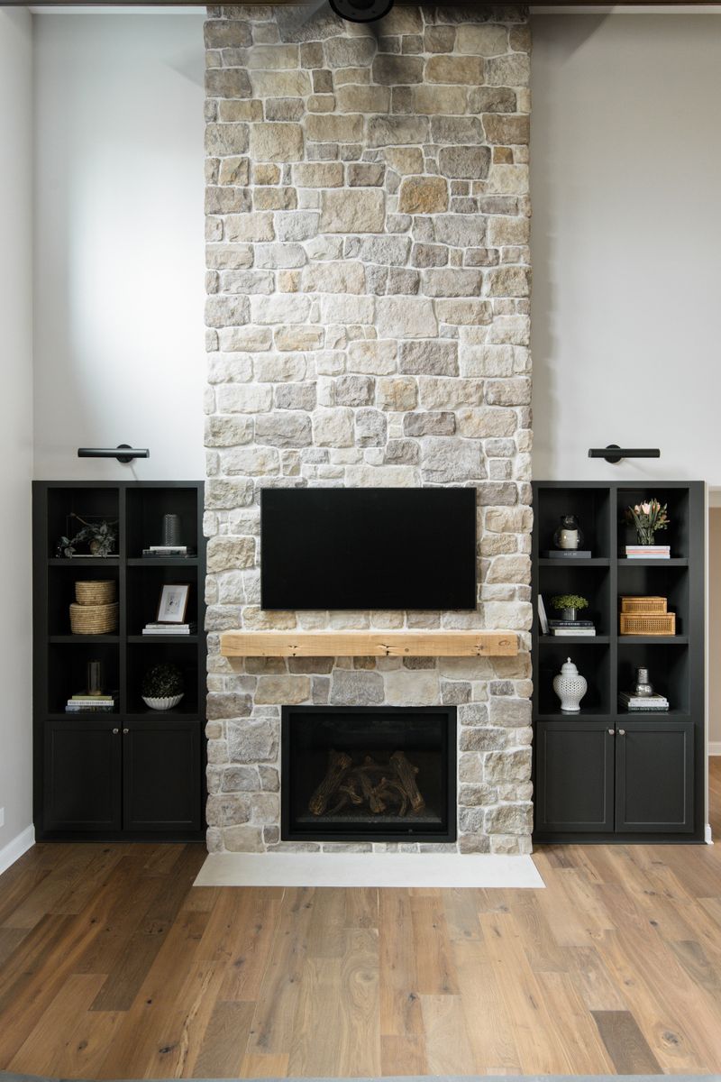 A living room in Van Meter with a stone double height, two-story fireplace and a raw wood mantel between black built-in shelves.