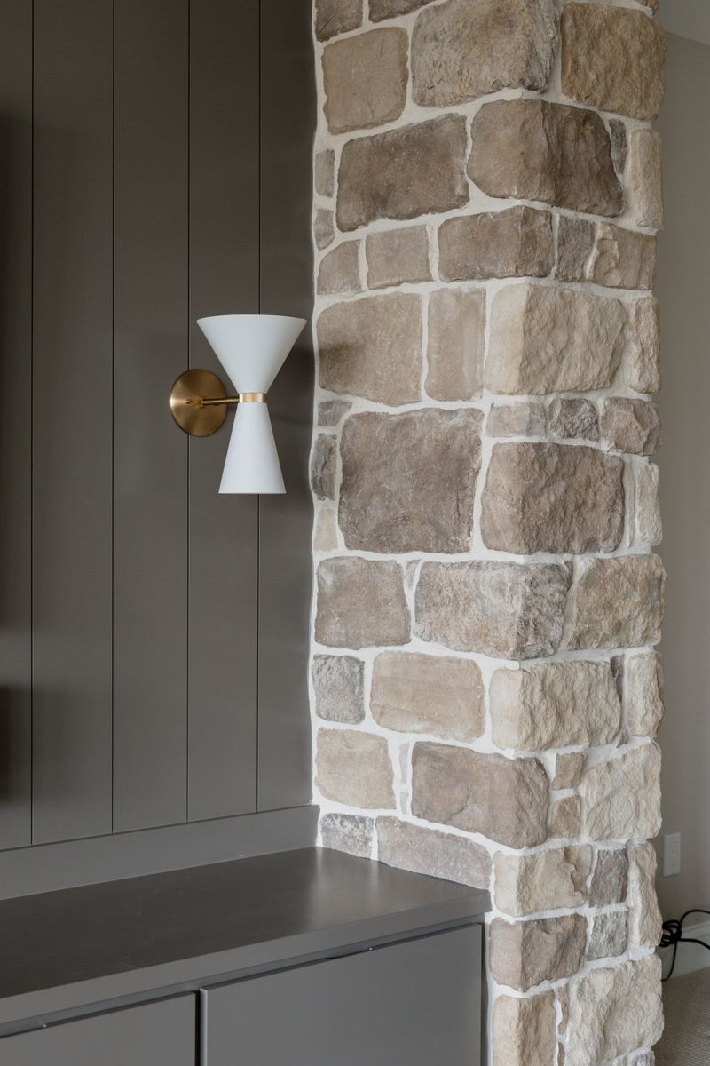 A mudroom in Van Meter with a white and brass sconce on gray paneling beside a stone wall.