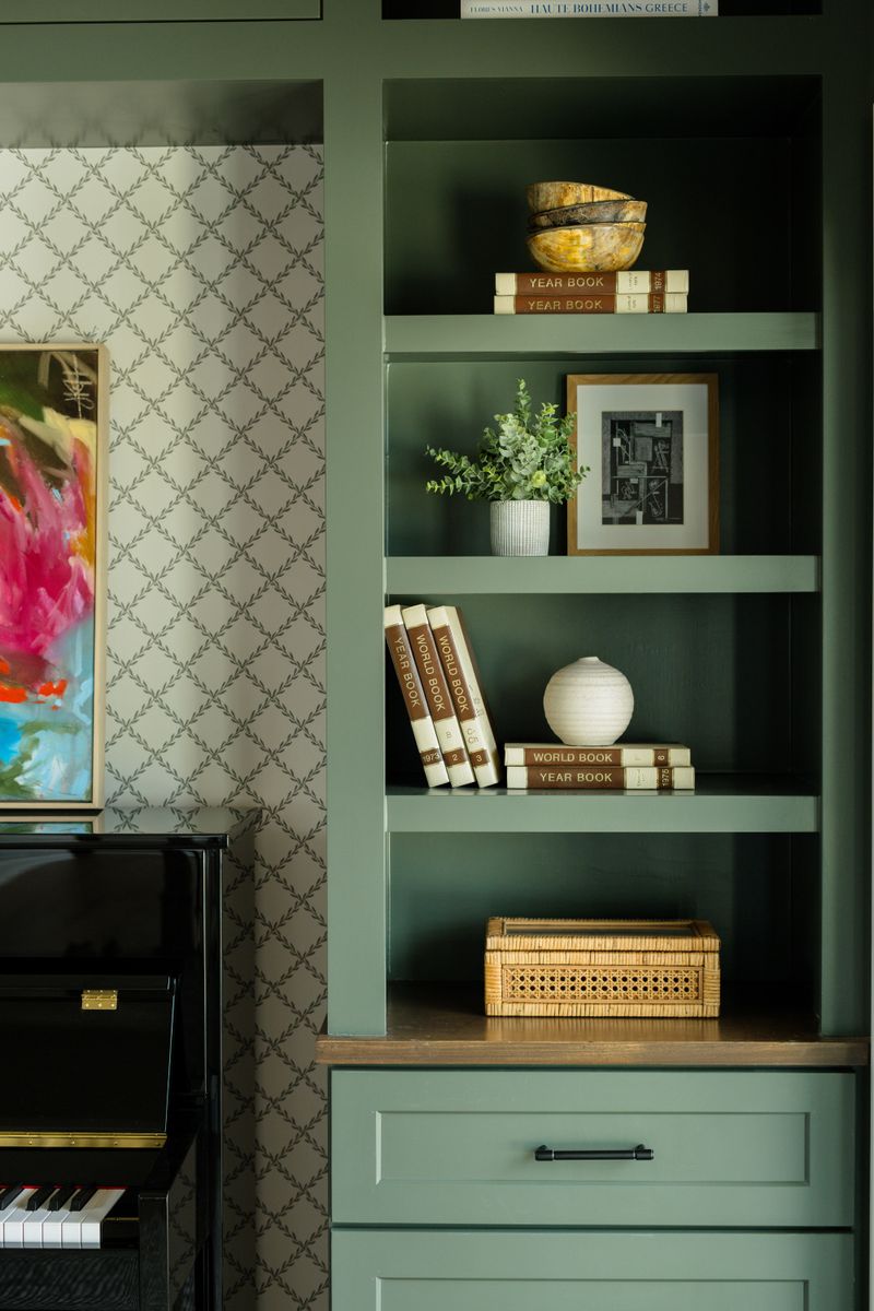 A music room in Van Meter with sage green built-in shelves beside a black upright piano and trellis wallpaper.