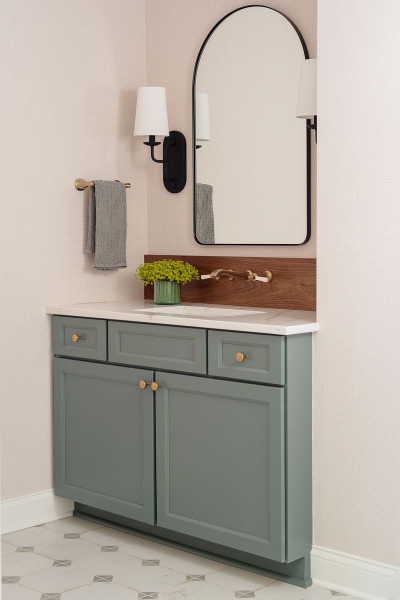 A powder bath in Van Meter with a green vanity and a walnut backsplash behind the brass wall faucet.