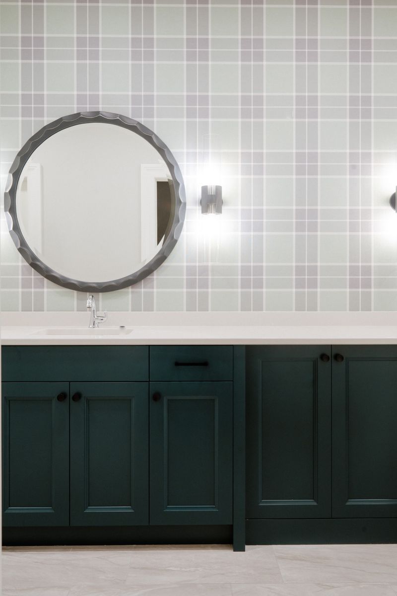 A bath in Urbandale with dark green vanity cabinets under gray plaid wallpaper and a round scalloped mirror.