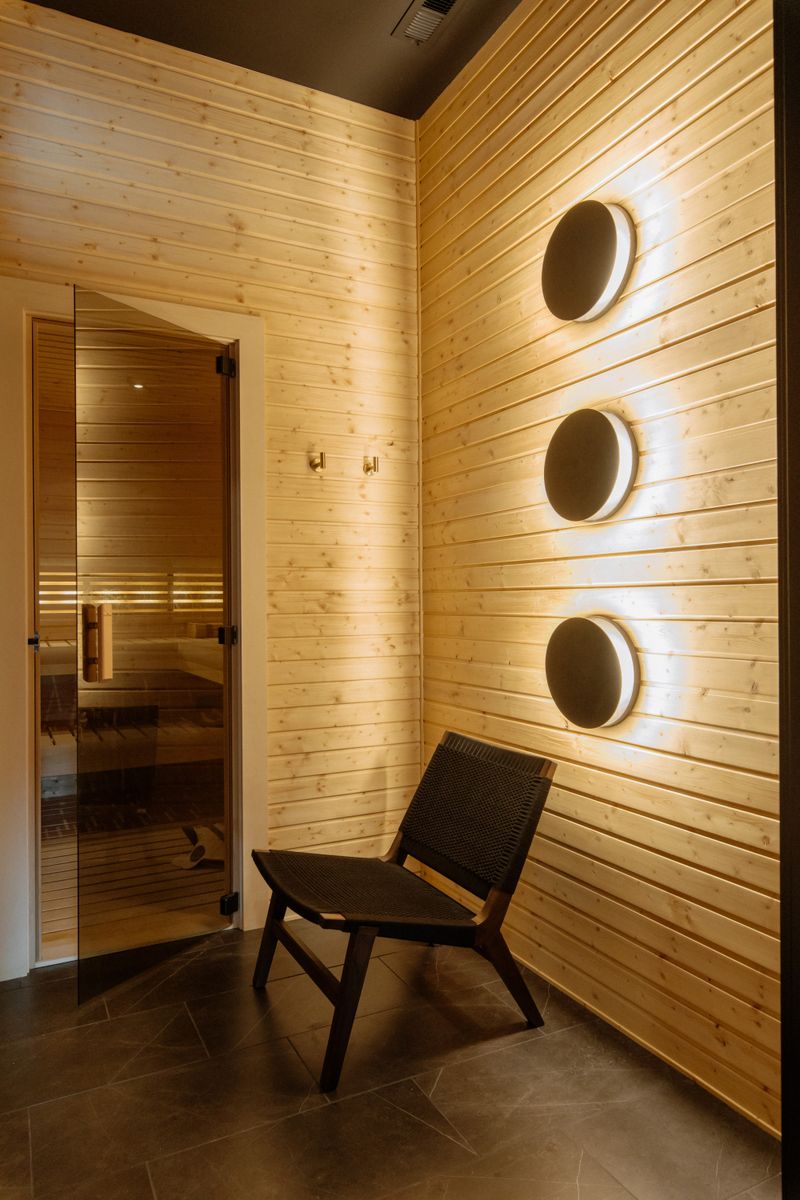 A cedar sauna in Urbandale with three round wall lights glowing on the plank wall beside a glass door.