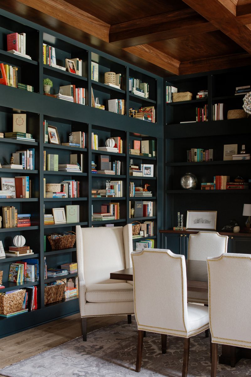 A home library and dining room combo in Urbandale with dark green built-in bookshelves and cream nailhead chairs at a wood table.