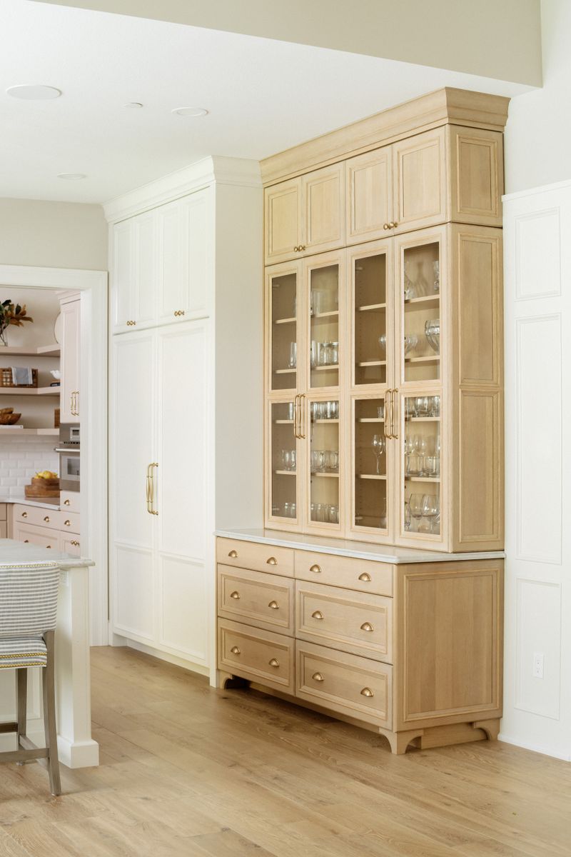 A kitchen in Urbandale with a white oak glass front hutch and brass cup pulls beside white paneled cabinets.