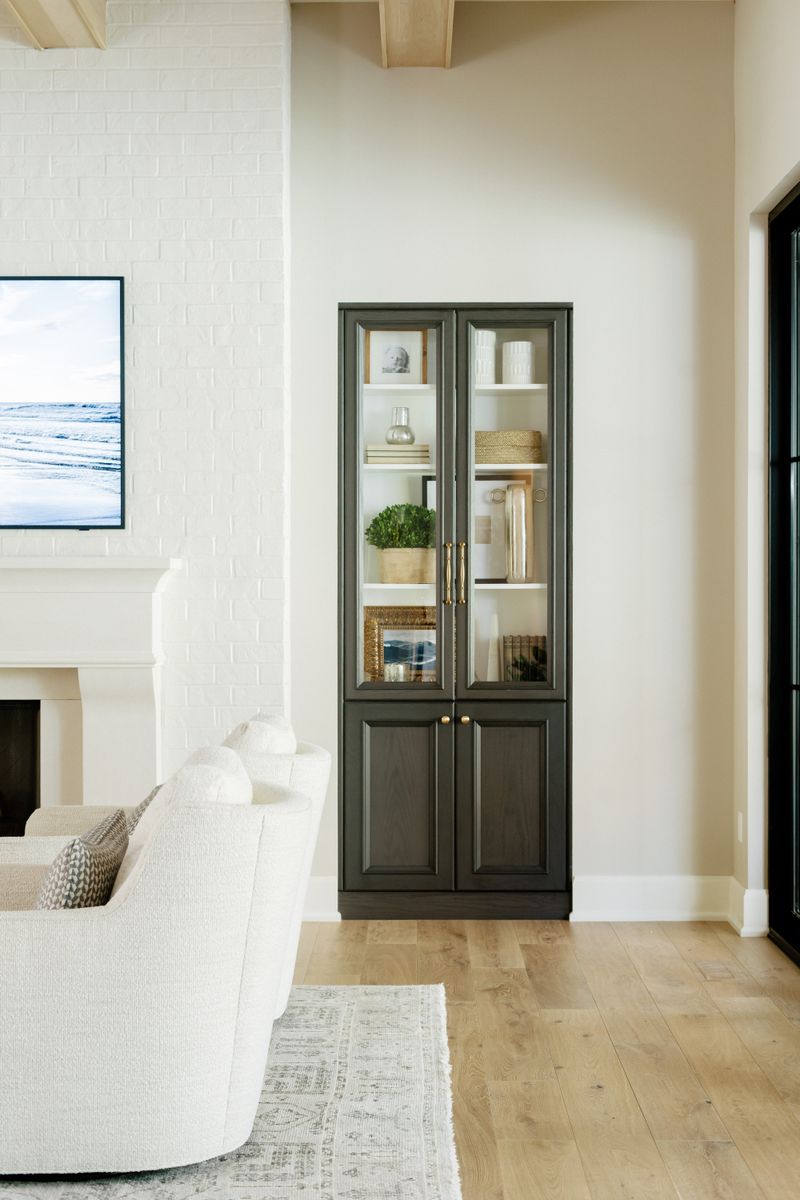 A living room in Urbandale with a dark green glass front cabinet and brass hardware beside a painted white brick fireplace.