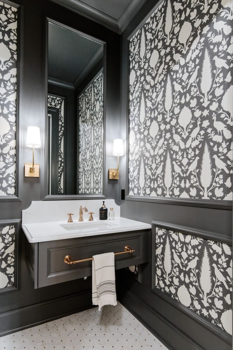 A powder room in Urbandale with black and white bird wallpaper above charcoal paneling and brass sconces.