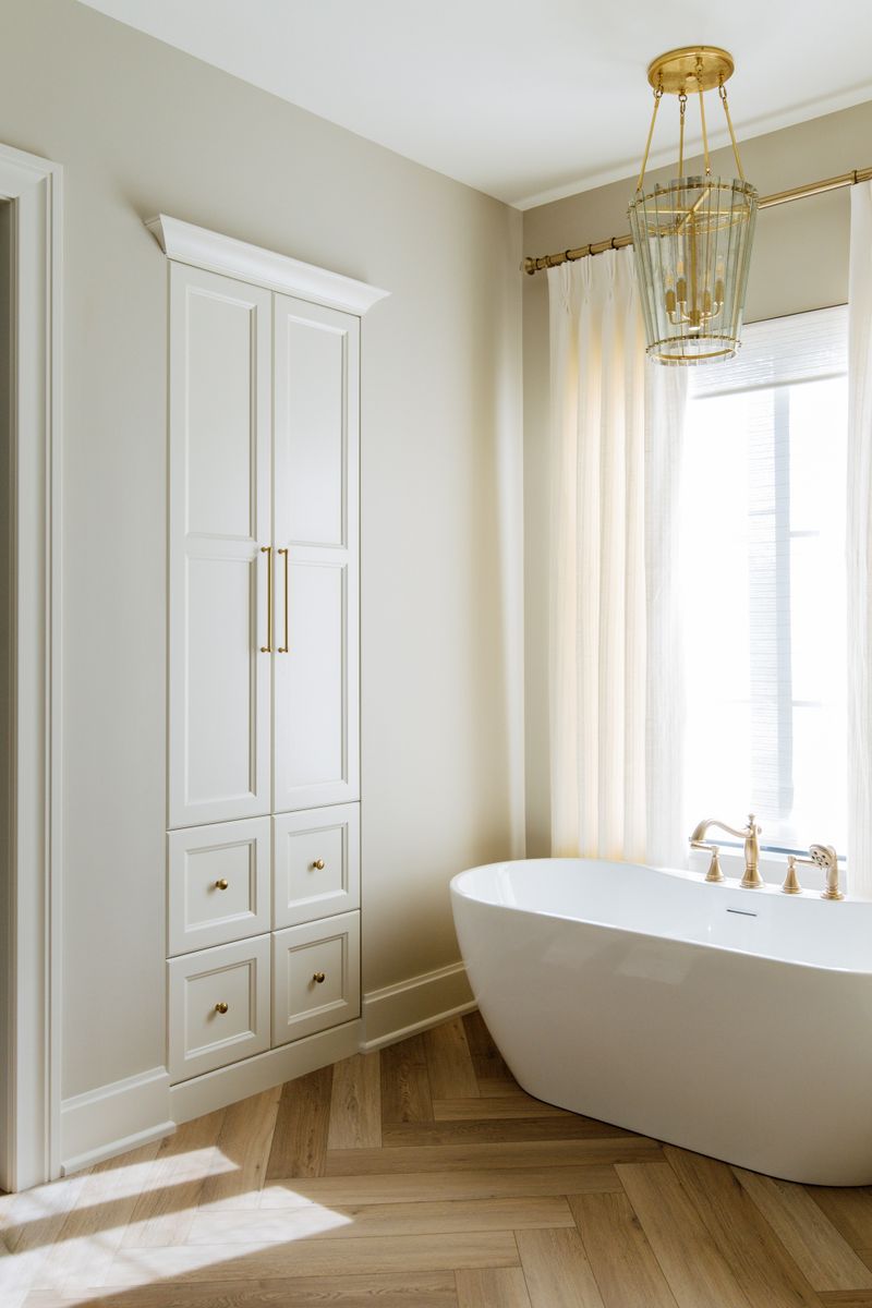 A primary bath in Urbandale with a freestanding white tub on herringbone wood floors and brass tub filler.