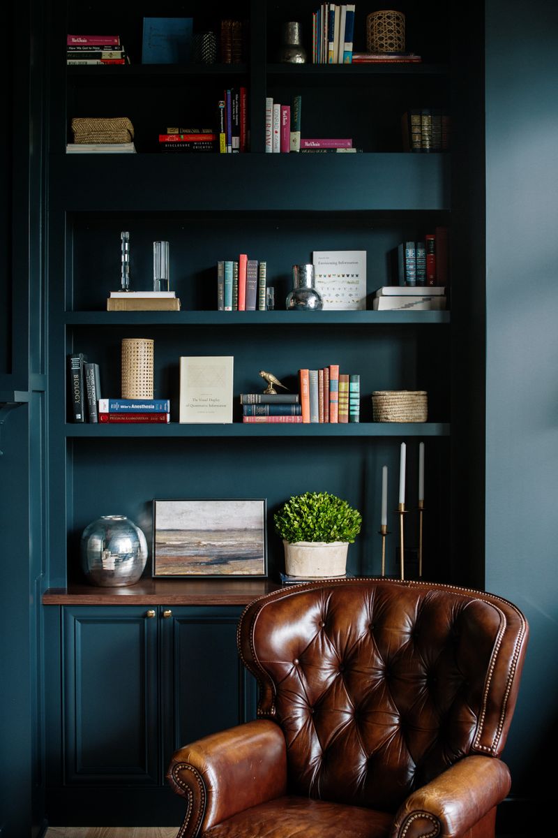 A study in Urbandale with deep teal built-in bookshelves behind a tufted brown leather armchair.
