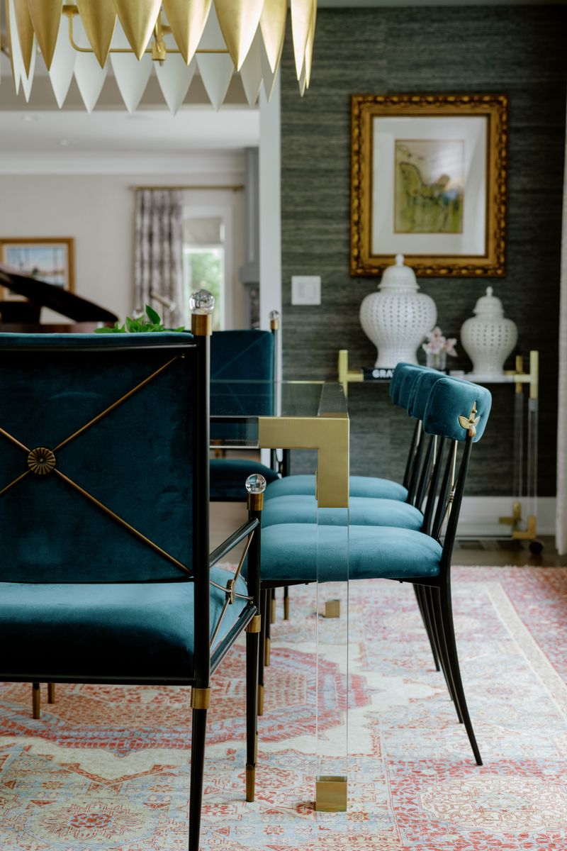 A Des Moines dining room with teal velvet chairs around a glass and brass table, set against dark grasscloth walls.