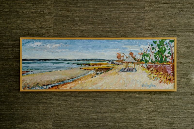 A framed landscape oil painting in a light wood frame hung on olive grasscloth wallpaper in Des Moines.