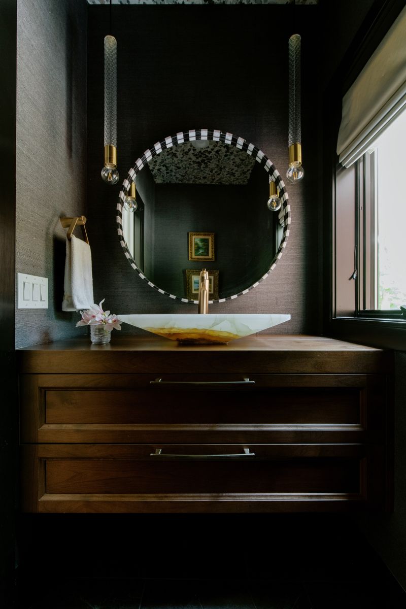 A dark powder room in Des Moines with a lit onyx vessel sink on a walnut vanity under a round mirror.