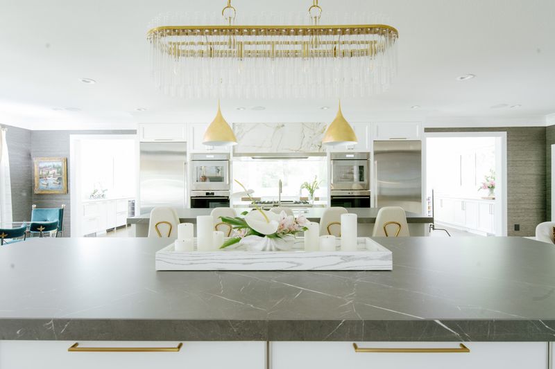 A white kitchen in Des Moines with a gray stone island and brass hardware under a glass chandelier.