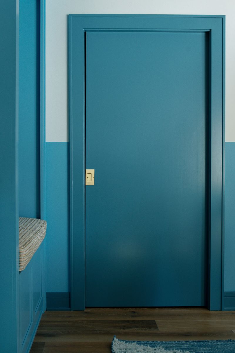 A teal mudroom door in Des Moines with a brass light switch plate and a built in bench beside it.