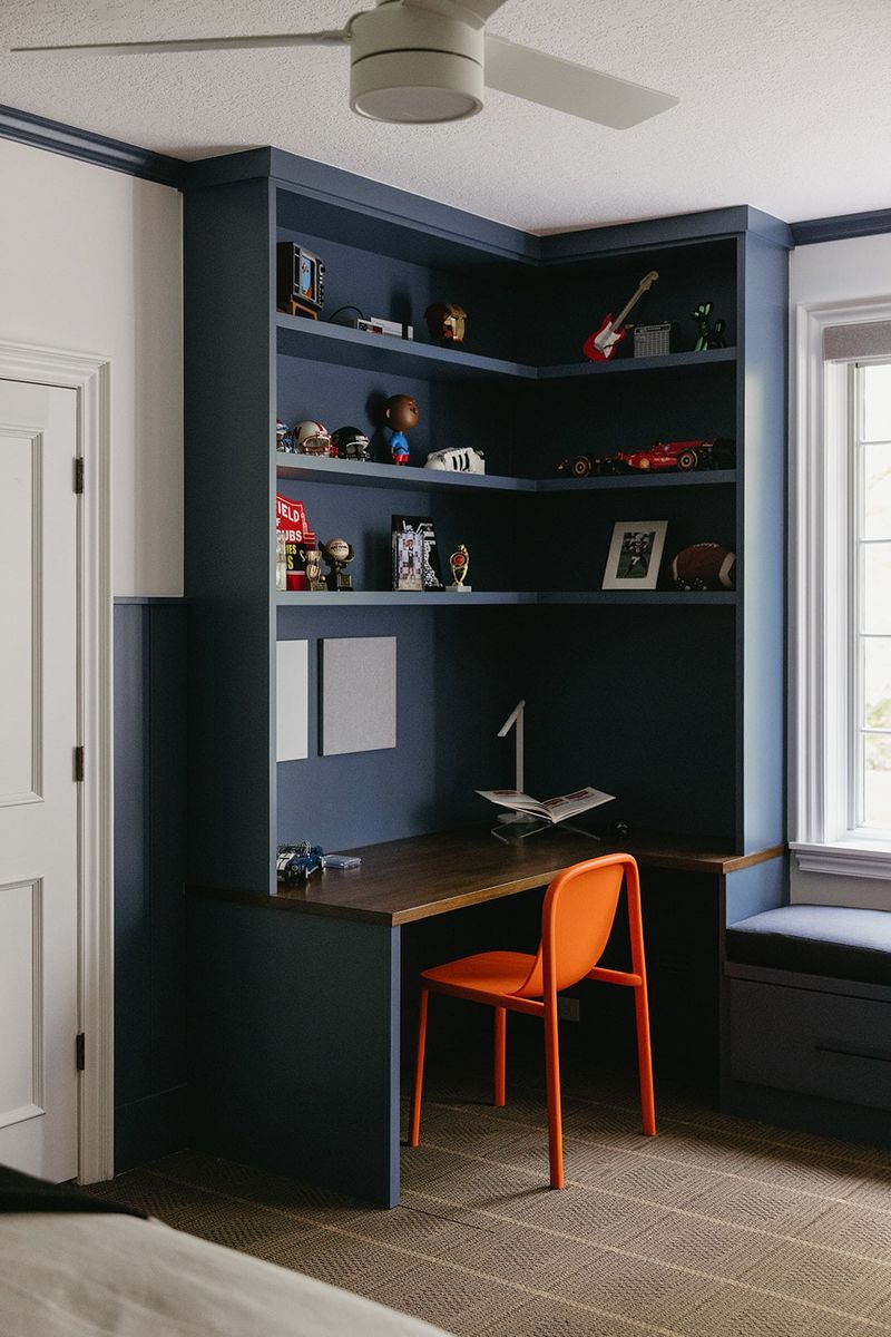 A navy blue built-in desk nook in a Clive bedroom with a walnut desktop and an orange chair.