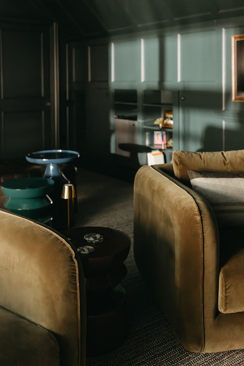 A moody green paneled living room in Clive with olive velvet sofas around a dark round coffee table.