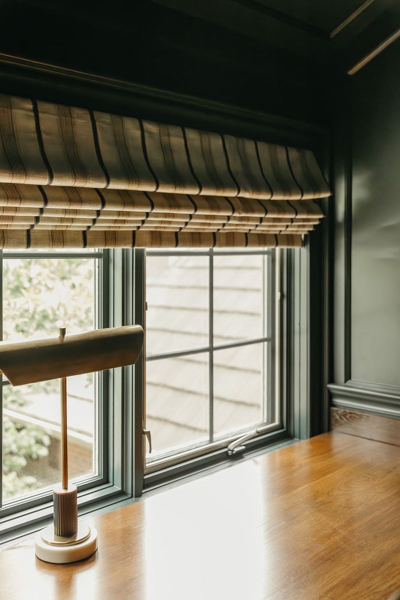 A green home office in Clive with a brass desk lamp on a wood desk under a woven roman shade.