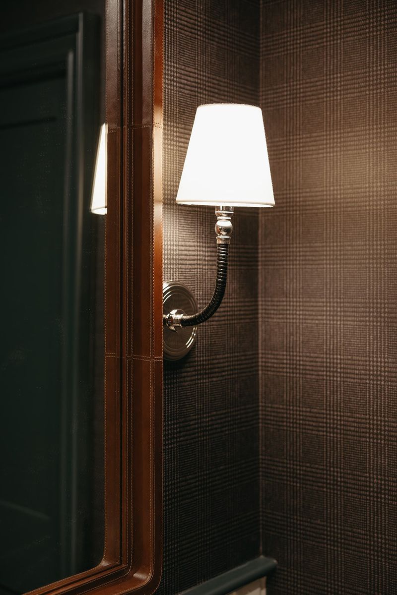A powder room in Clive with a nickel sconce on brown plaid wallpaper, beside a stitched leather mirror frame.