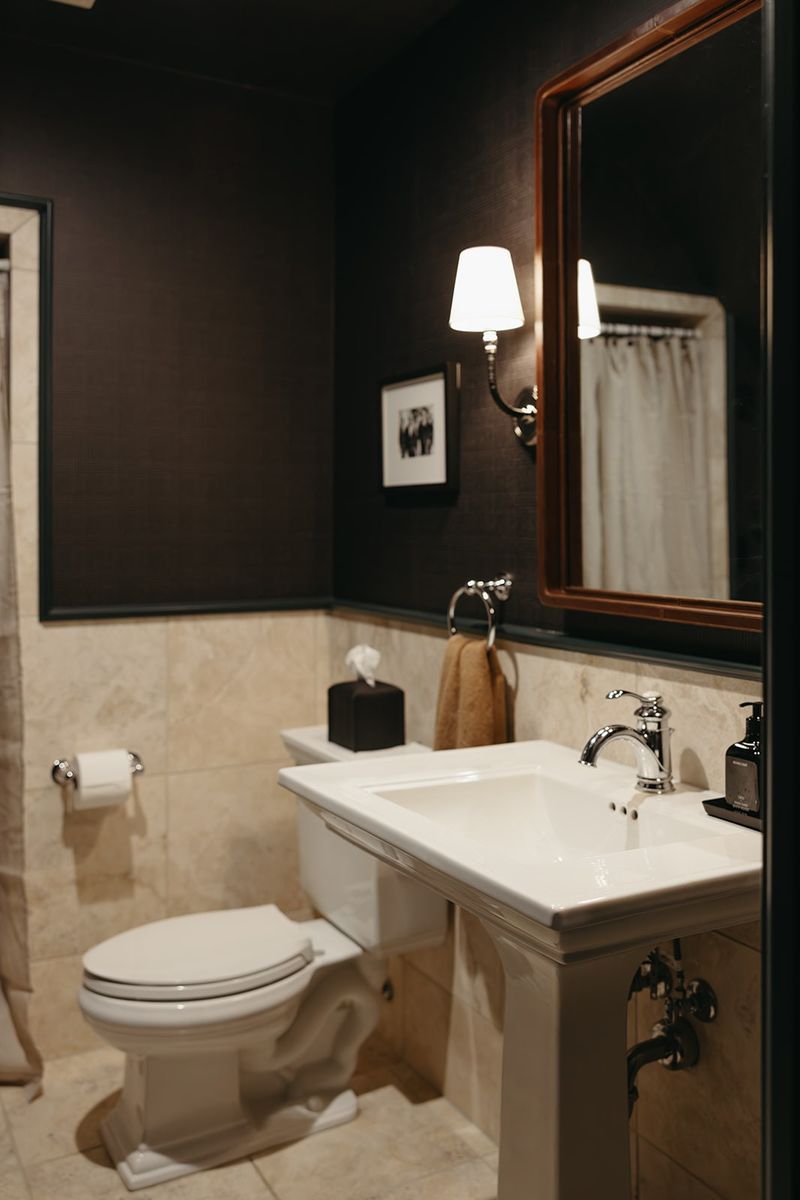 A powder room in Clive with dark brown walls above cream marble tile and a white pedestal sink.