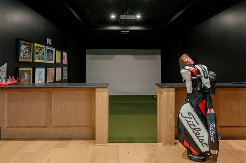 A golf simulator room in West Des Moines with a black ceiling and a white oak bar counter beside the putting turf.