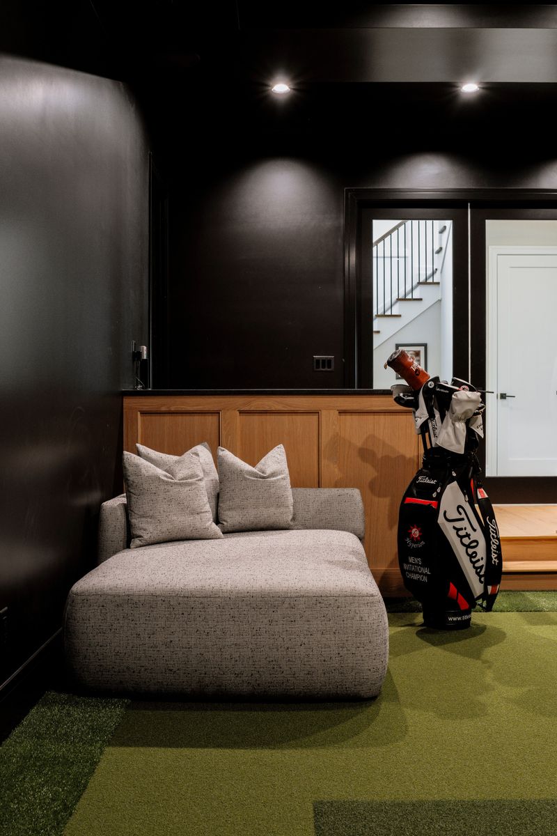 A golf simulator room in West Des Moines with black walls above white oak paneling and green turf flooring.