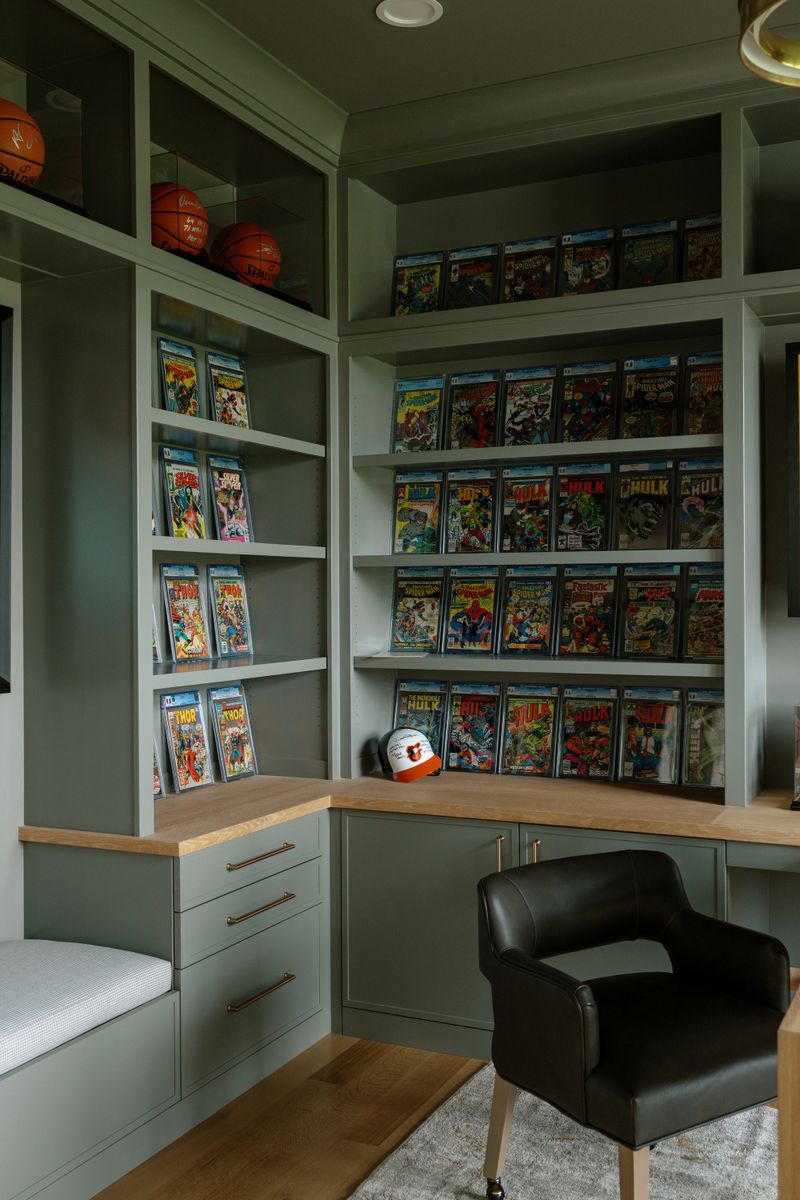 A home office in West Des Moines with sage green built-in shelves displaying comic books above a white oak desk.