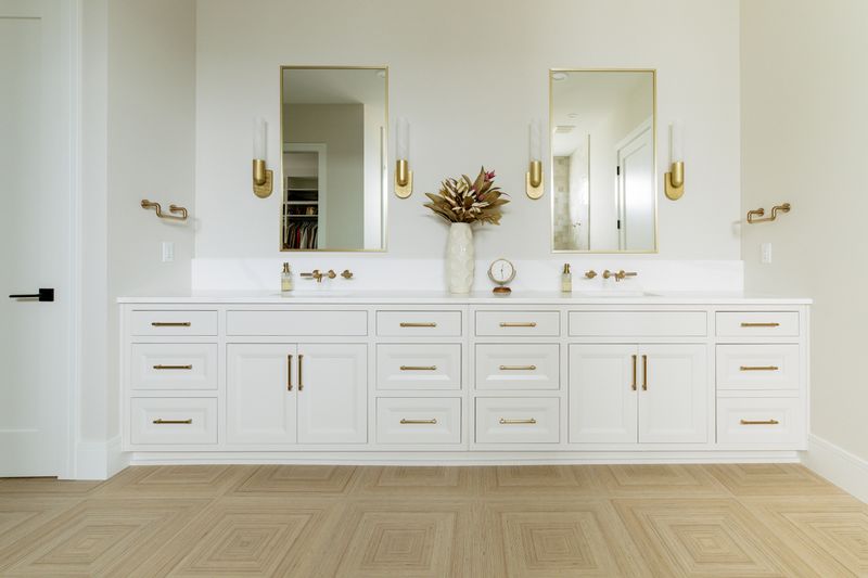 A primary bath in West Des Moines with a long white vanity and brass hardware over light herringbone wood floors.