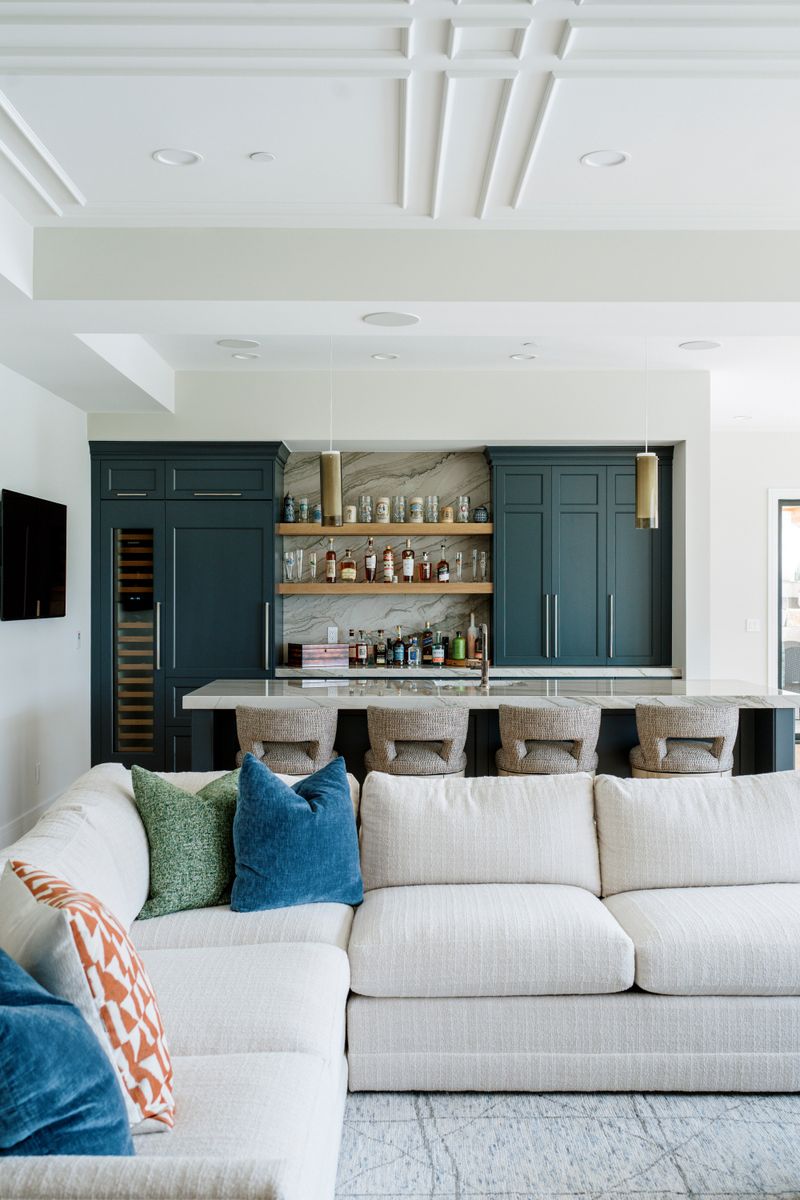 A wet bar in West Des Moines with deep teal cabinets and a marble backsplash behind oak floating shelves.