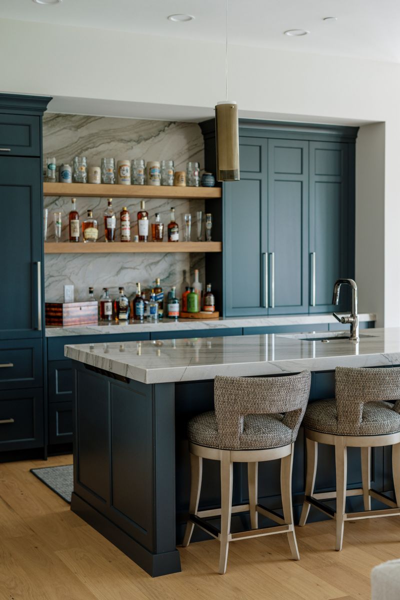 A navy wet bar in West Des Moines with white oak floating shelves against a veined marble backsplash.