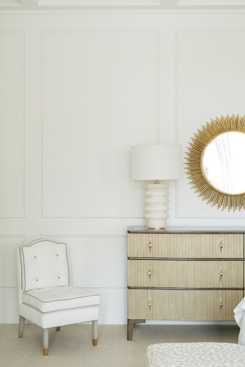 A cream bedroom in Des Moines with a gold sunburst mirror above a fluted dresser, set against paneled walls.