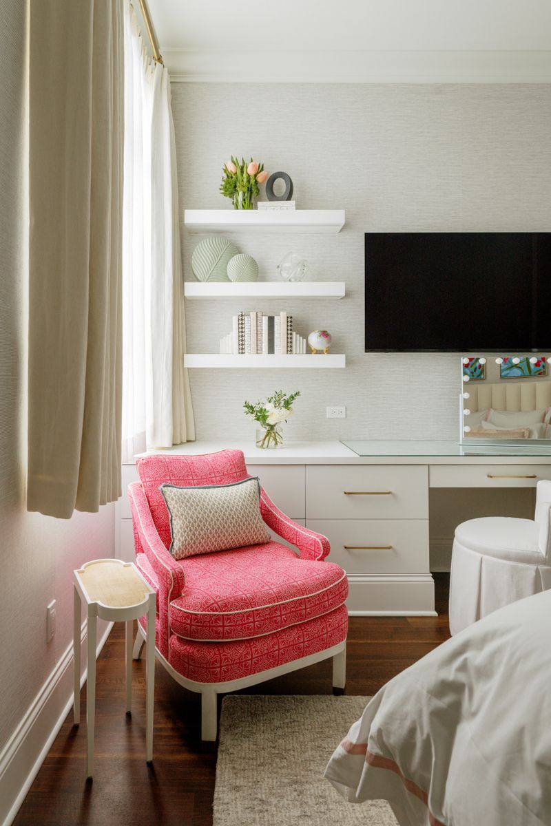 A bedroom in Des Moines with a pink patterned armchair beside a built-in white vanity and floating shelves.