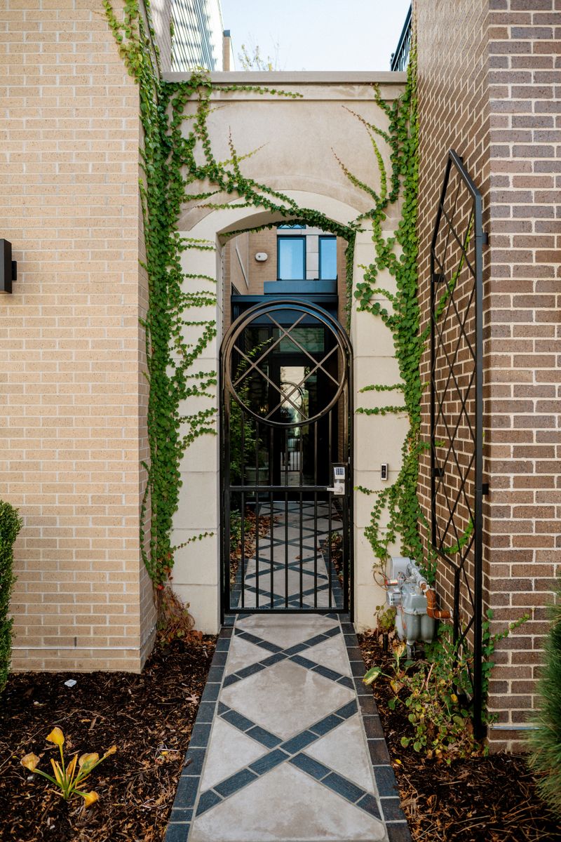 A Des Moines entry with a black iron gate under a vine-covered stone arch, over a diamond-patterned stone walkway.