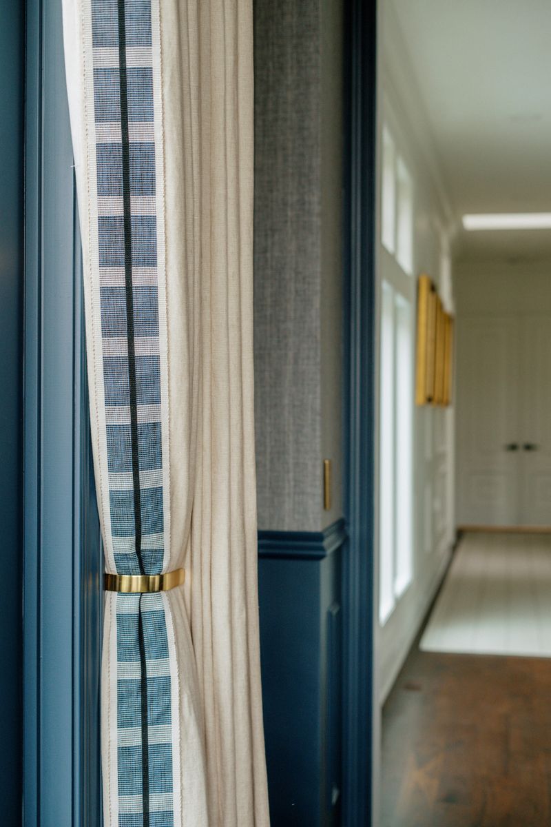 A navy hallway in Des Moines with a brass tieback holding a linen curtain trimmed in blue check.