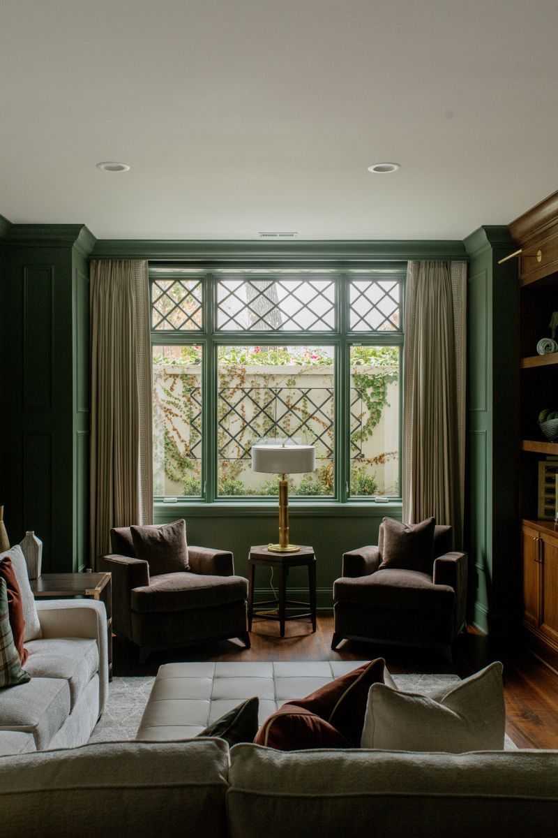 A dark green paneled living room in Des Moines with a brass lamp between two velvet armchairs at the window.
