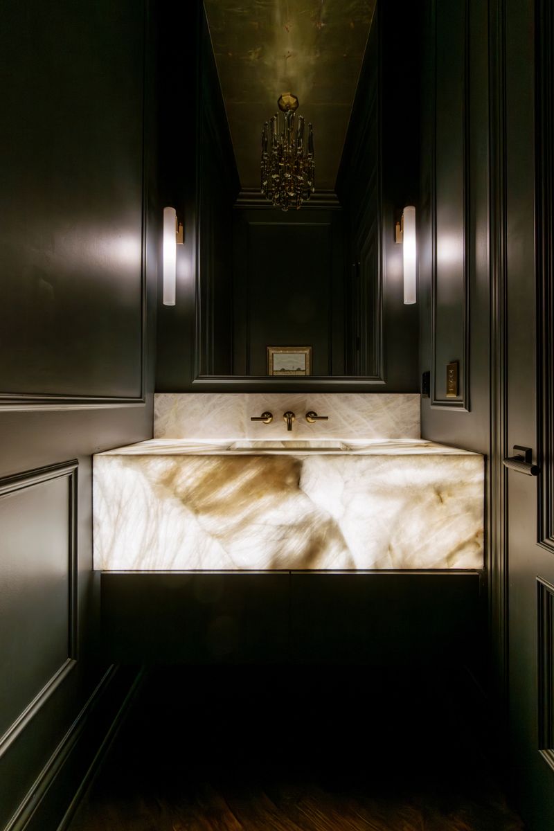 A dark grey powder room in Des Moines with a backlit onyx vanity and a brass wall mounted faucet.