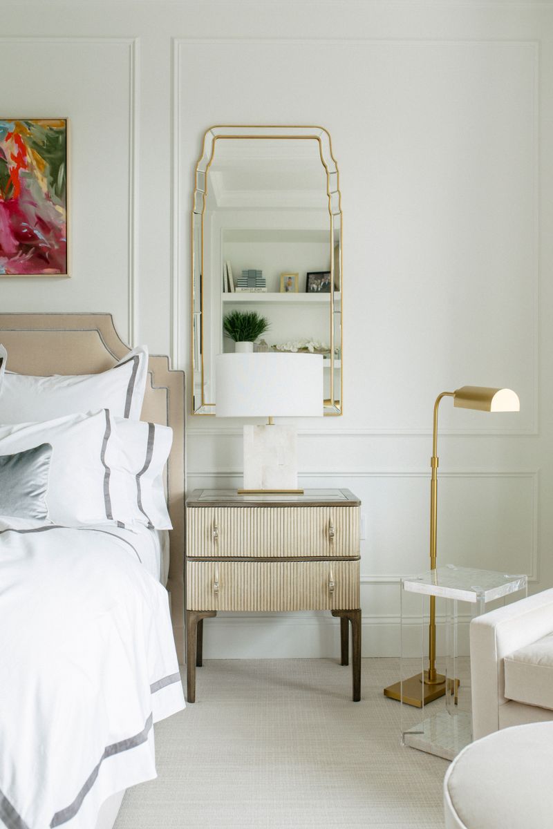 A soft white primary bedroom in Des Moines with a fluted gold nightstand under a tall brass mirror.
